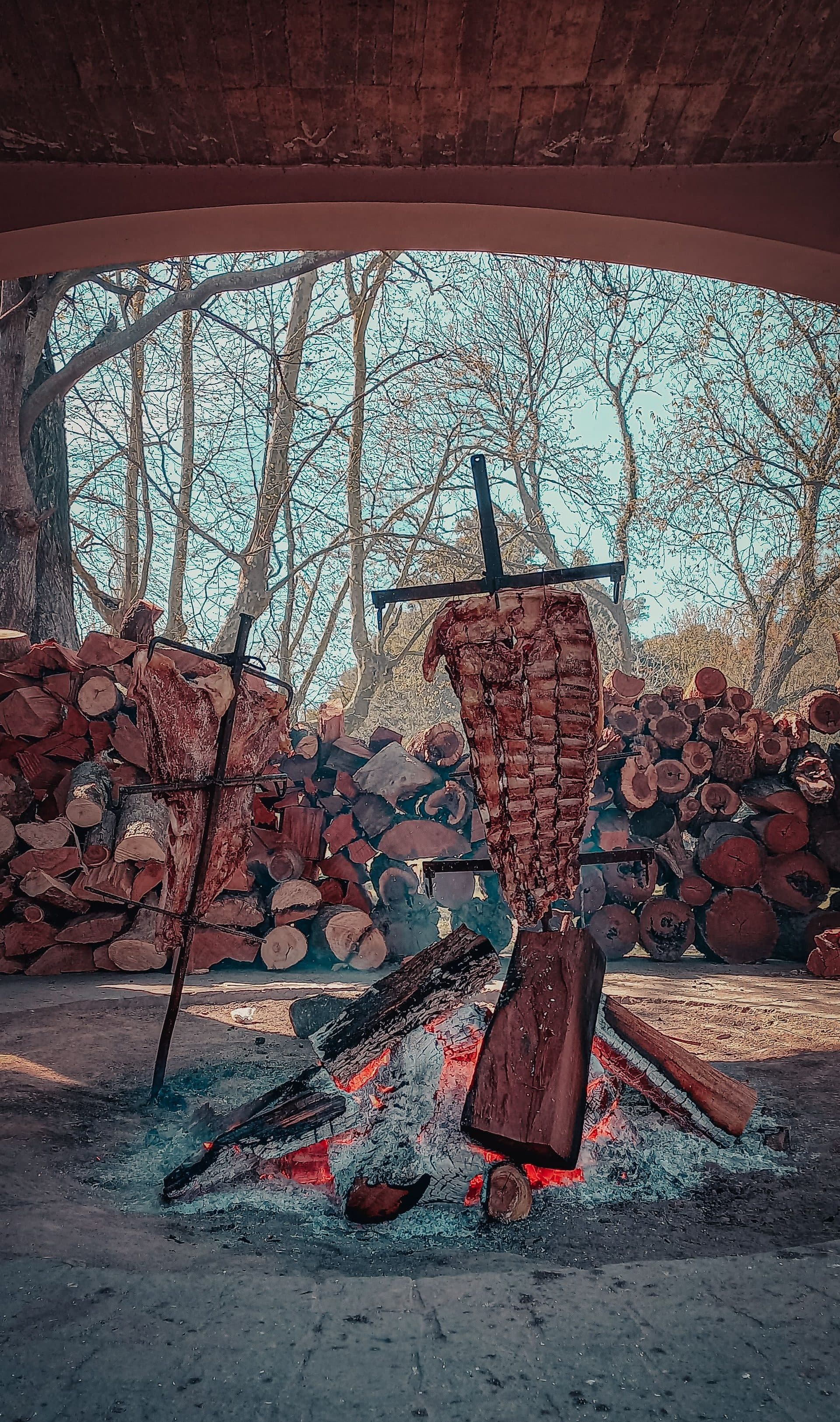 Rinder-Asado am Kreuzspieß (Asador) über offener Feuerstelle, im Hintergrund gestapeltes Brennholz — Symbolbild